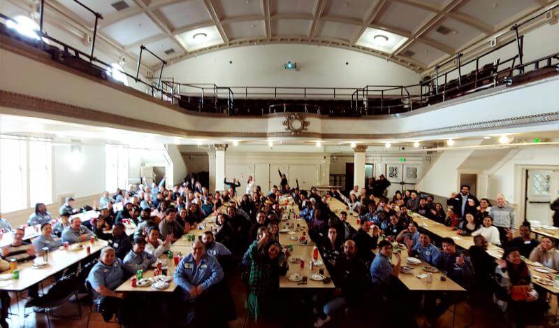 over 100 people sit together in a huge auditorium eating lunch 