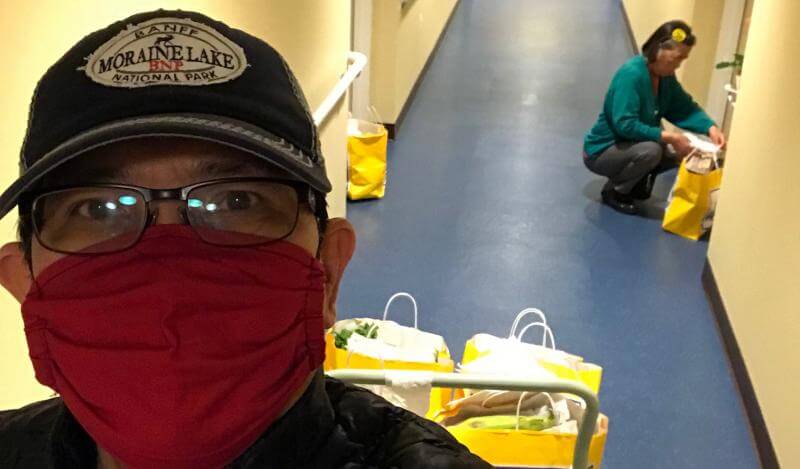 A staffmember in a mask takes a selfie with bags of food in background, another person helps set them at doors in a hallway