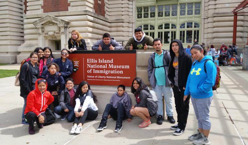 a multi-racial group of teens pose in front of Ellis Island