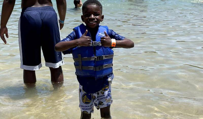 A young black kid wearing a life-jacket shows two thumbs up at a lake