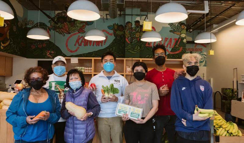 A diverse group of staff hold various food items in a pantry with a mural reading "Kain Na" and "Let's Eat!"