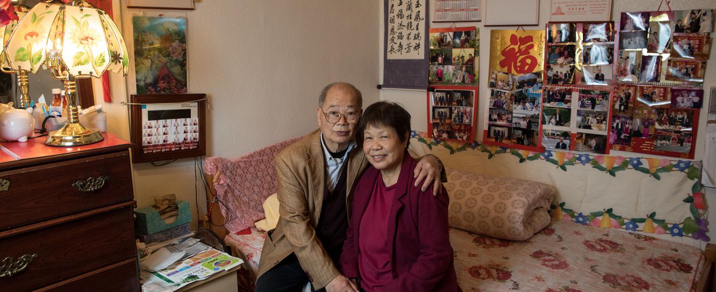 Senior Chinese couple sits on a patterned bed with many photos and posters in background