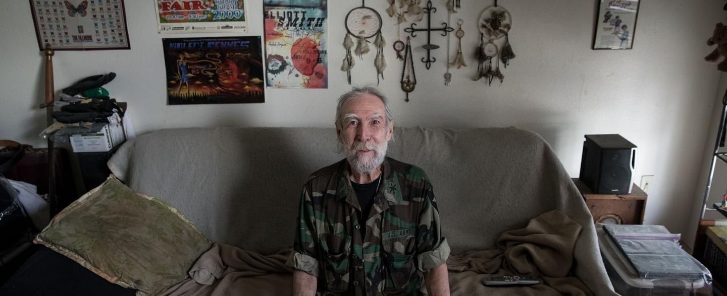 Senior male sits at his couch with posters and dream-catcher on back wall