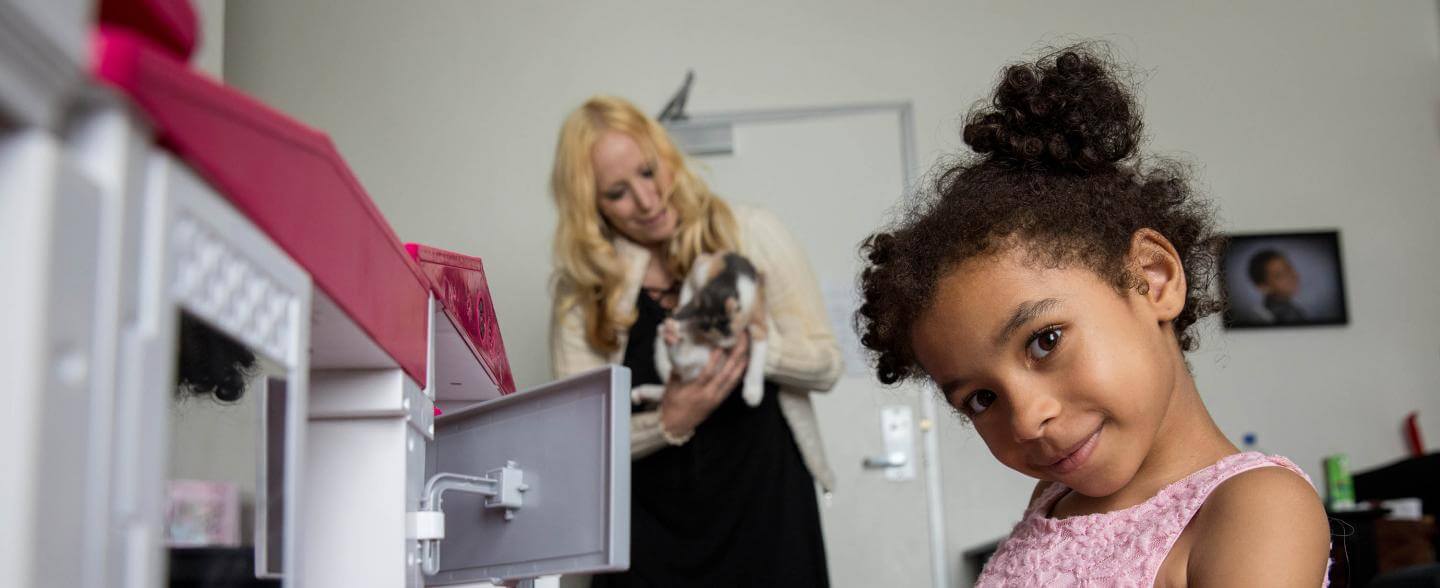 A young black girl plays with her toys while her white mom stands in the background