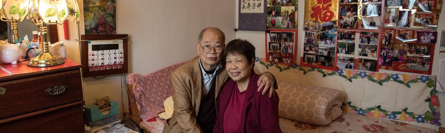 Senior Chinese couple sits on a patterned bed with many photos and posters in background