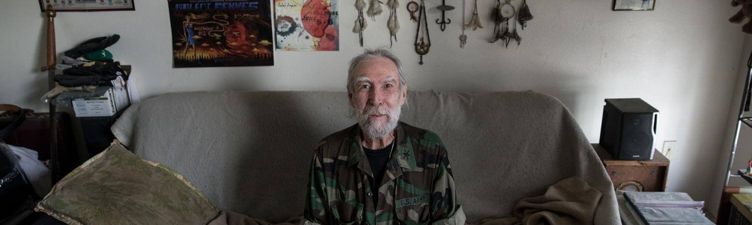 Senior male sits at his couch with posters and dream-catcher on back wall