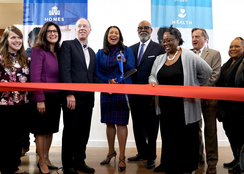 A multi-racial group including Mayor London Breed pose and cut a red ribbon