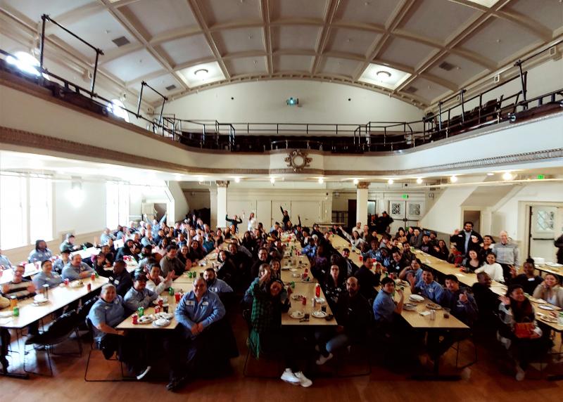 over 100 people sit together in a huge auditorium eating lunch 