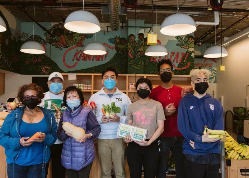 A diverse group of staff hold various food items in a pantry with a mural reading "Kain Na" and "Let's Eat!"