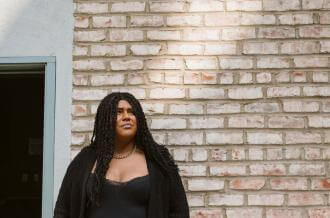A woman in a black shirt stands in front of a brick wall looking upwards 