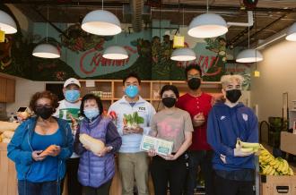 A diverse group of staff hold various food items in a pantry with a mural reading "Kain Na" and "Let's Eat!"